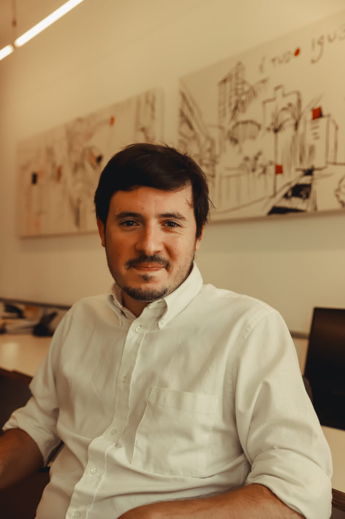 Foto de Francisco Brito Cruz, homem branco, de bigodes, de cabelos curtos e pretos. Veste uma blusa de mangas compridas branca. Ao fundo, há dois quadros com ilustrações de cartões-postais da Cidade de São Paulo. A foto é de Bianca Moreira.