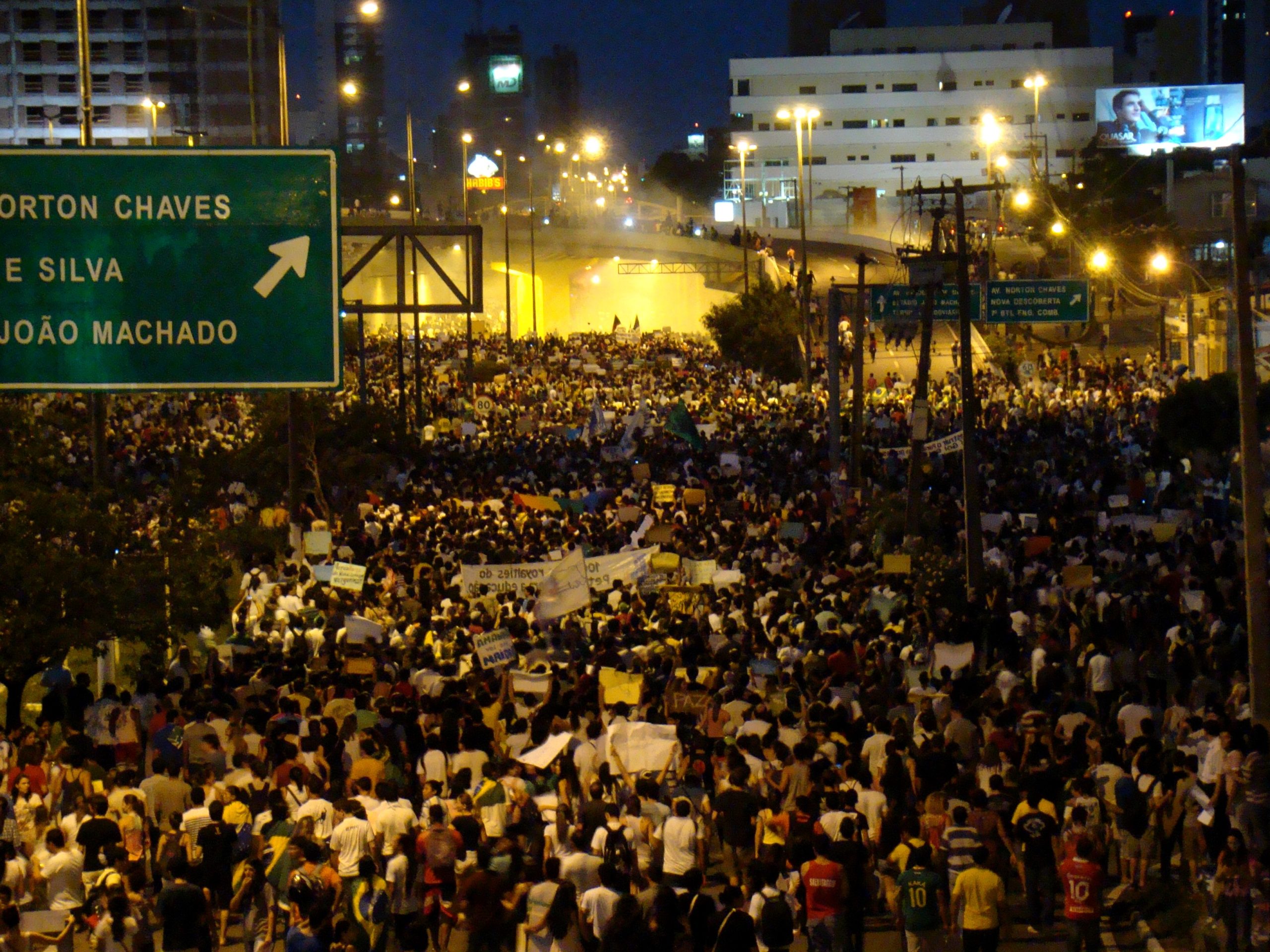 Foto de protestantes sentados em uma avenida. Algumas pessoas seguram cartazes.
