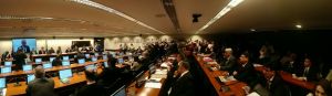 Foto panorâmica do interior da Câmara dos Deputados. Os deputados estão de terno sentados em suas bancadas, com uma tela em frente.
