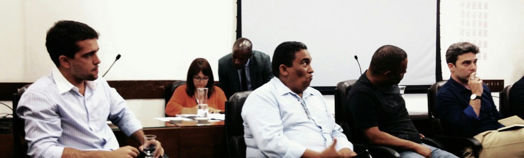 Foto de pessoas sentadas em uma mesa de debate. Na primeira fileira, estão sentados quatro homens. Na mesa atrás, está sentada uma mulher e um homem está agachado ao lado dela, conversando.