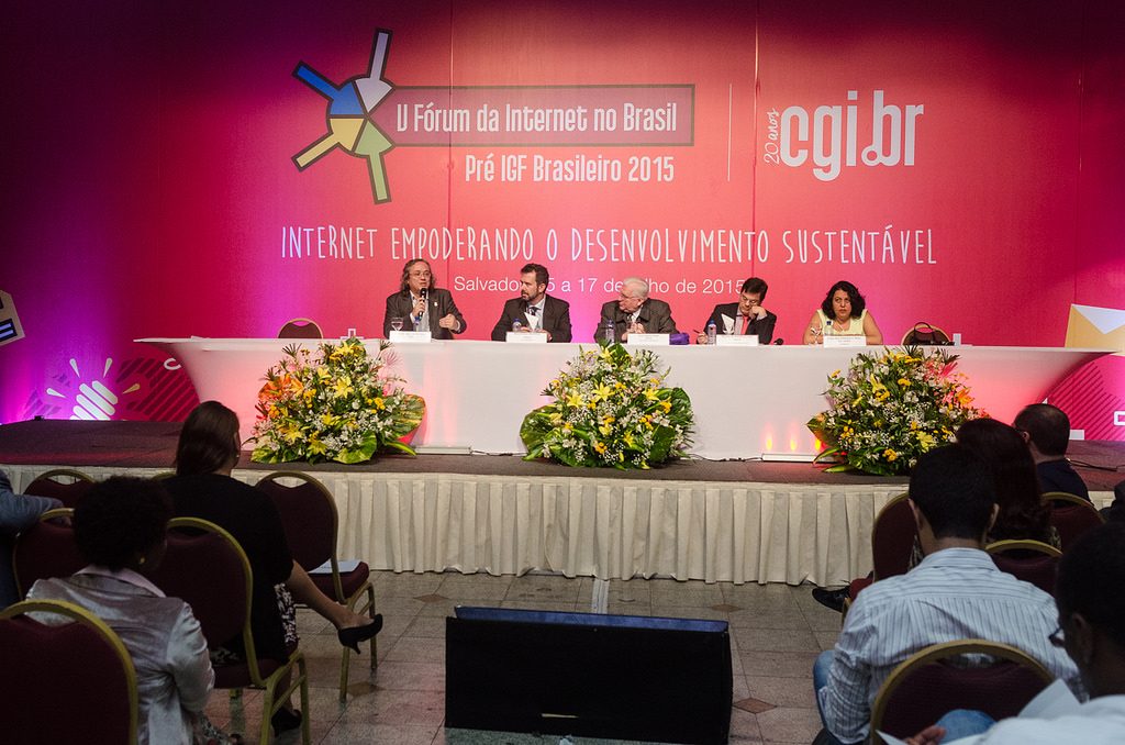 Foto do Fórum da Internet, onde se encontram cinco pessoas sentadas na mesa diretora do evento, em frente a um painel rosa com os logotipos do evento "V Fórum da Internet no Brasil: Pré IGF Brasileiro 2015" e do cgi.br e o título do evento "Internet empoderando o desenvolvimento sustentável:". Na frente da mesa, estão dispostos 3 ornamentos de flores e o público sentado em fileiras.