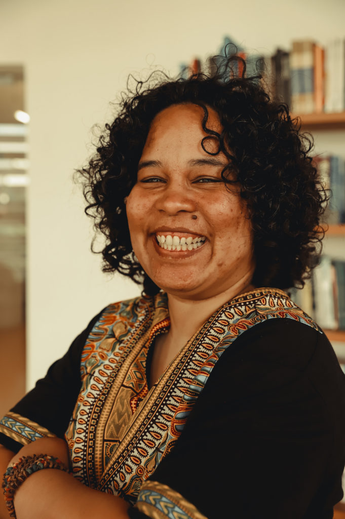 Imagem de Alessandra Gomes, mulher negra, de cabelos cacheados curtos. Aparece na foto sorrindo, vestindo blusa preta com detalhes coloridos. Foto de Bianca Moreira/InternetLab.