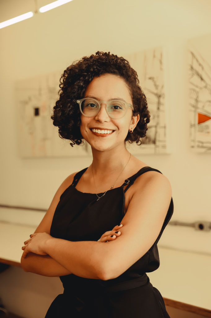 Foto de Ester Borges, mulher negra, de cabelos cacheados e curtos, acima dos ombros. Porta óculos de grau, aparece sorrindo e de braços cruzados, vestida com blusa preta de alças. No fundo da imagem há um quadro branco. Os créditos da foto são de Bianca Moreira/InternetLab.