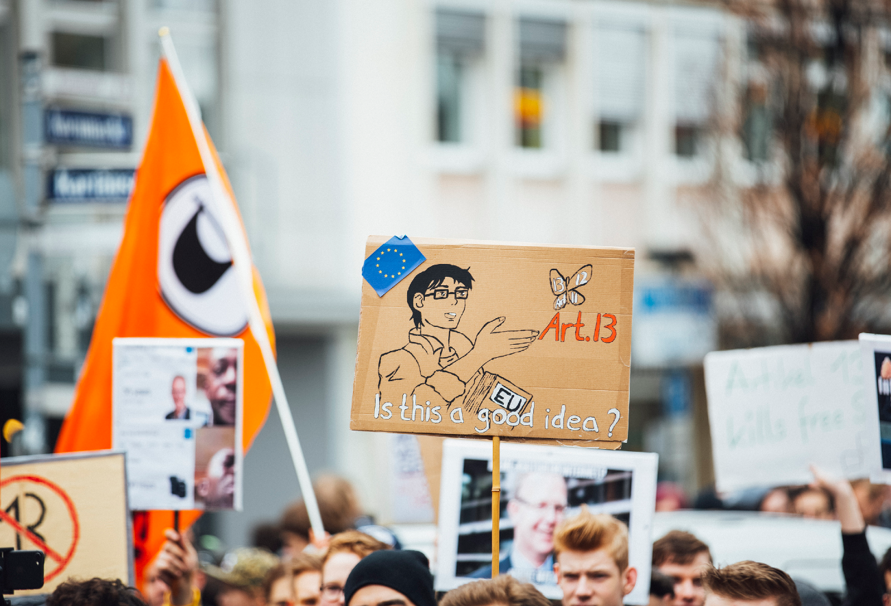 Foto de uma manifestação exibindo placas e bandeiras carregadas pelos manifestantes com destaque em uma placa com o desenho de um personagem de anime apontando para um borboleta, fazendo referência ao meme is this a Pigeon? e o texto: Art. 13 is this a good idea?