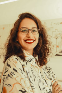 Foto de Clarice Tavares, mulher negra, de cabelos cacheados e curtos. Aparece sorrindo e de braços cruzados, vestida com blusa de mangas curtas branca com grafismos coloridos. No fundo da imagem há um quadro branco. A foto é de Bianca Moreira/InternetLab.