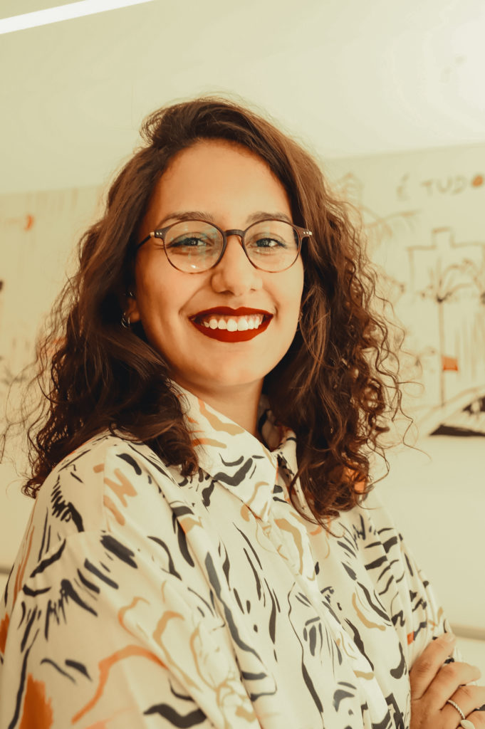 Foto de Clarice Tavares, mulher negra, de cabelos cacheados e curtos. Aparece sorrindo e de braços cruzados, vestida com blusa de mangas curtas branca com grafismos coloridos. No fundo da imagem há um quadro branco. A foto é de Bianca Moreira/InternetLab.