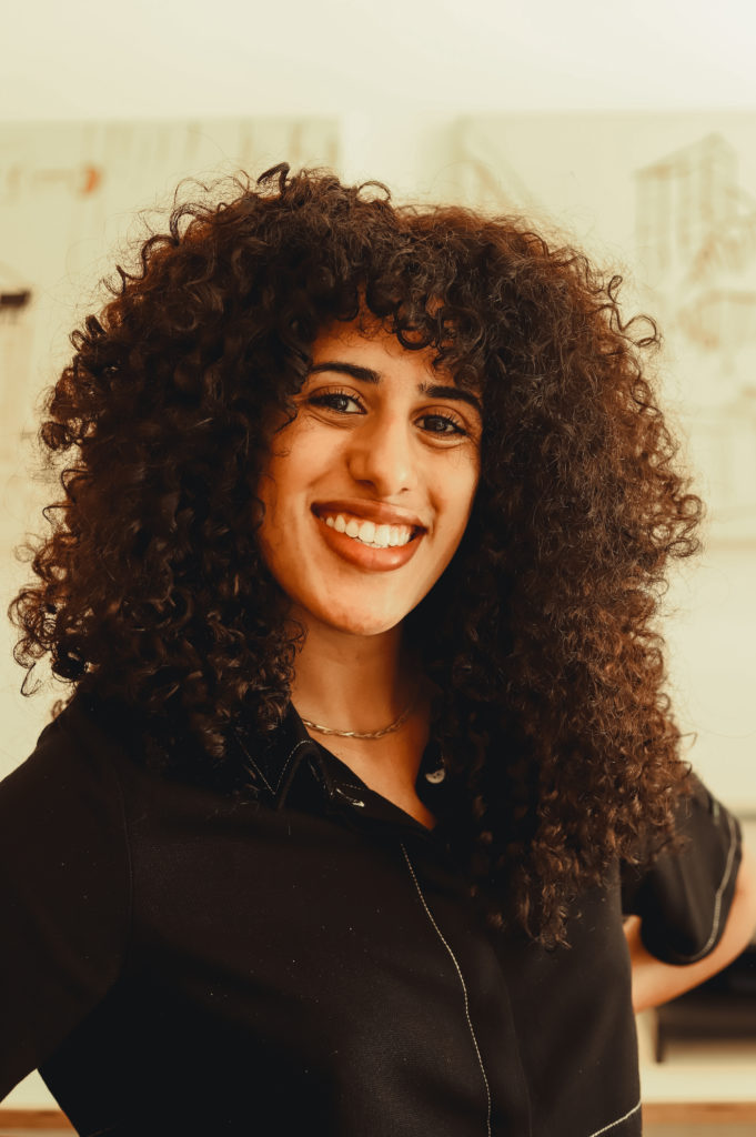 Foto de Catharina Pereira, mulher negra de cabelos cacheados e castanhos. Na imagem, aparece sorrindo, vestida com uma blusa preta. Foto de Bianca Moreira/InternetLab