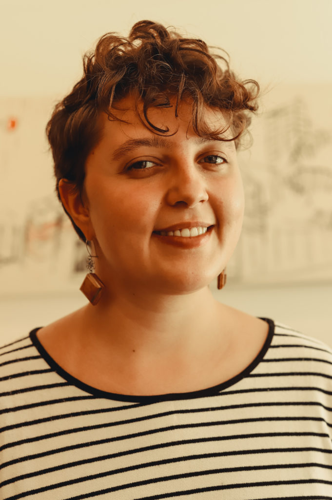 Foto de Jade Becari, mulher branca, de cabelos castanhos claros e curtos. Aparece na foto sorrindo, vestida com uma listrada nas cores branca e preta. Créditos da foto para Bianca Moreira/InternetLab.