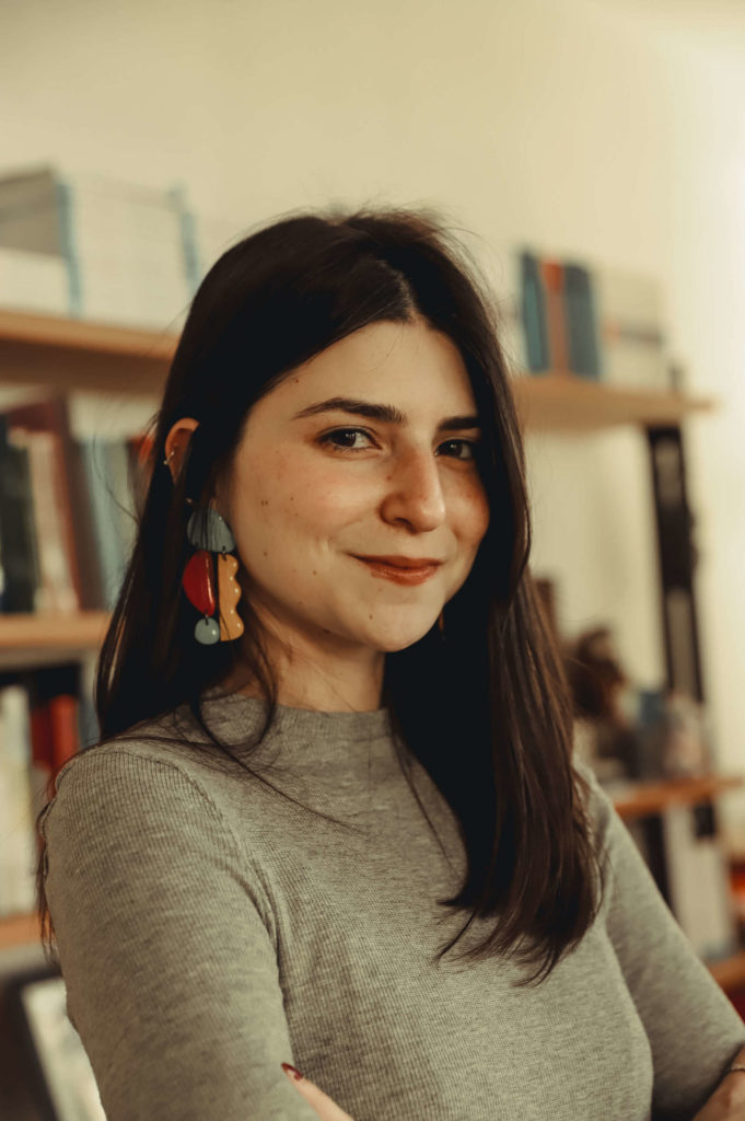 Foto de Juliana Fonteles, mulher branca, de cabelos castanhos e longos. Aparece de braços cruzados, sorrindo, com batom escuro nos lábios, vestida de manga longa na cor cinza e brincos coloridos. No fundo da imagem há uma estante de livros. Créditos da imagem para Bianca Moreira/InternetLab.