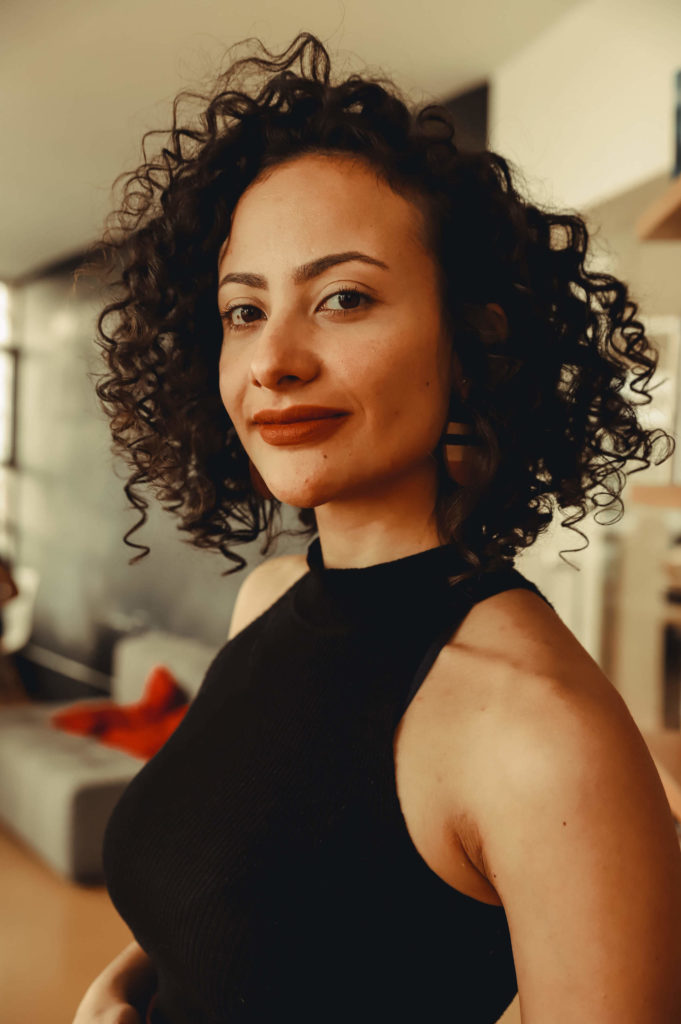Foto de Karina Oliveira, mulher negra, de cabelos curtos e castanhos. Aparece sorrindo, com batom marrom e vestida com uma blusa regata de gola média preta. No fundo da imagem há uma parede escura e um sofá. Créditos da imagem para Bianca Moreira/InternetLab.