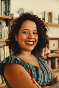 Foto de Stephanie Lima, mulher negra de cabelos cacheados e escuros. Na imagem, aparece sorrindo, vestida com uma blusa regata colorida e está com os braços cruzados. Ao fundo, há uma estante de livros. Créditos da imagem para Bianca Moreira/InternetLab.