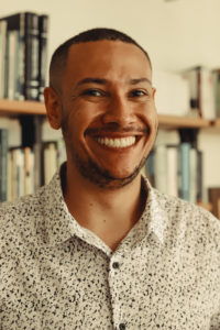 Foto de Luiz Fernando Sabino, homem negro, de barba e cabelos curtos. Ele veste blusa branca com detalhes pretos e botões na frente. e está sorrindo. Créditos para Bianca Moreira/InternetLab.