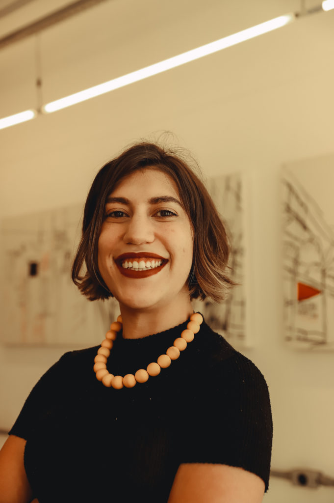 Imagem de Alice de Perdigão Lana, mulher banca de cabelos curtos e lisos. Aparece na foto sorrindo, com um colar de bolinhas e vestida com uma blusa de mangas curtas preta. Ao fundo, aparece um quadro.