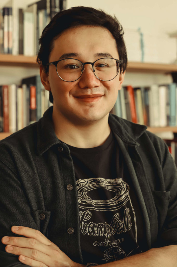 Foto de André Houang, homem asiático de cabelos pretos. Aparece sorrindo de óculos de grau e roupas pretas. No fundo, aparecem livros. Foto de Bianca Moreira/InternetLab.